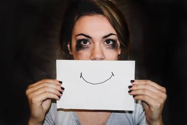 Traumatherapie, ein Lächeln ist nur eine Maske, um die Traurigkeit zu verbergen. Bild zeigt Frau, die vor ihr trauriges Gesicht ein aufgesetztes Lächeln auf Papier vorhält.