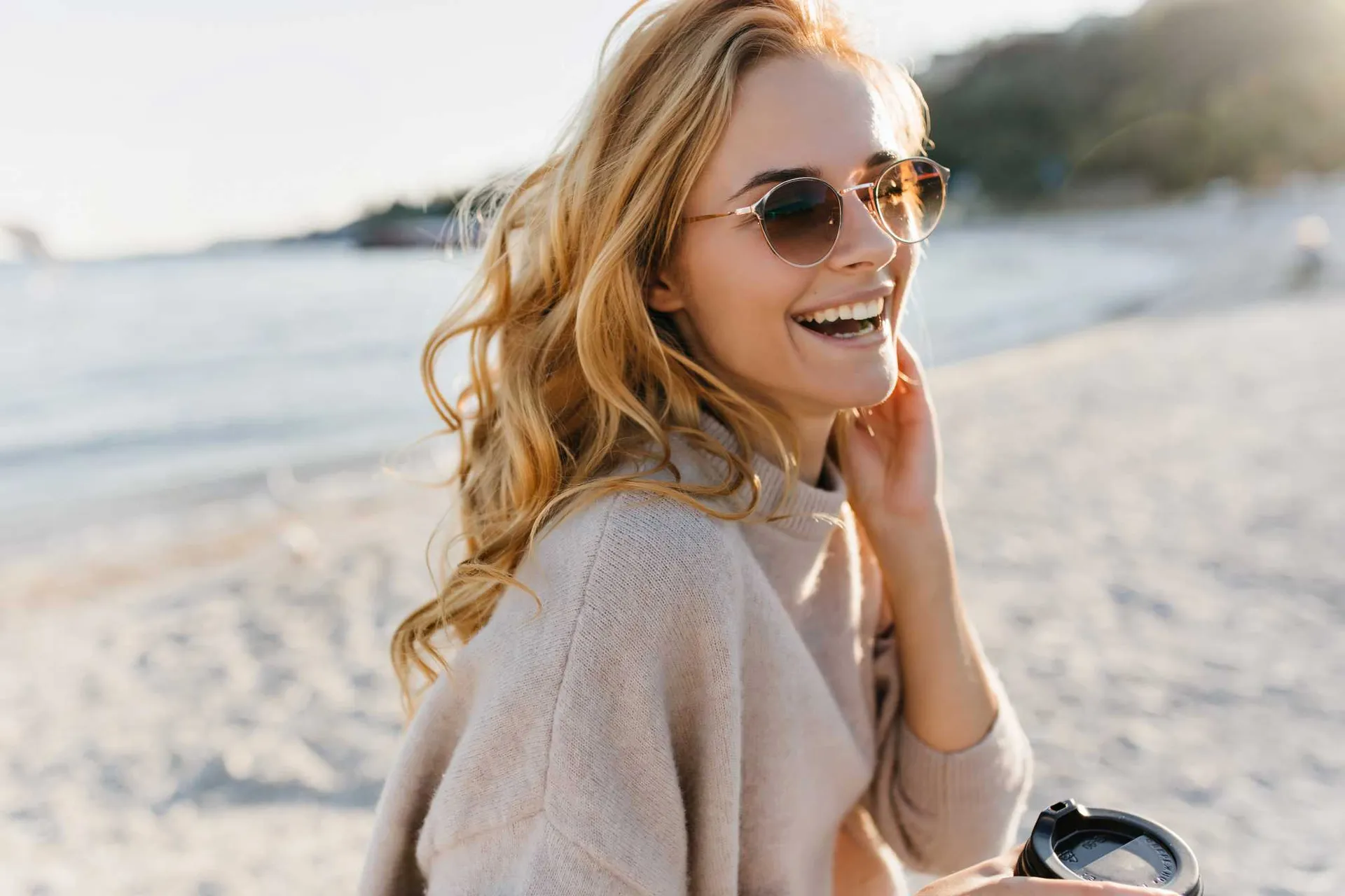 cute-woman-genuinely-laughs-relaxing-in-beach-blinde-woman-in-glasses-and-sweater-holds-cup-of-coffee-1